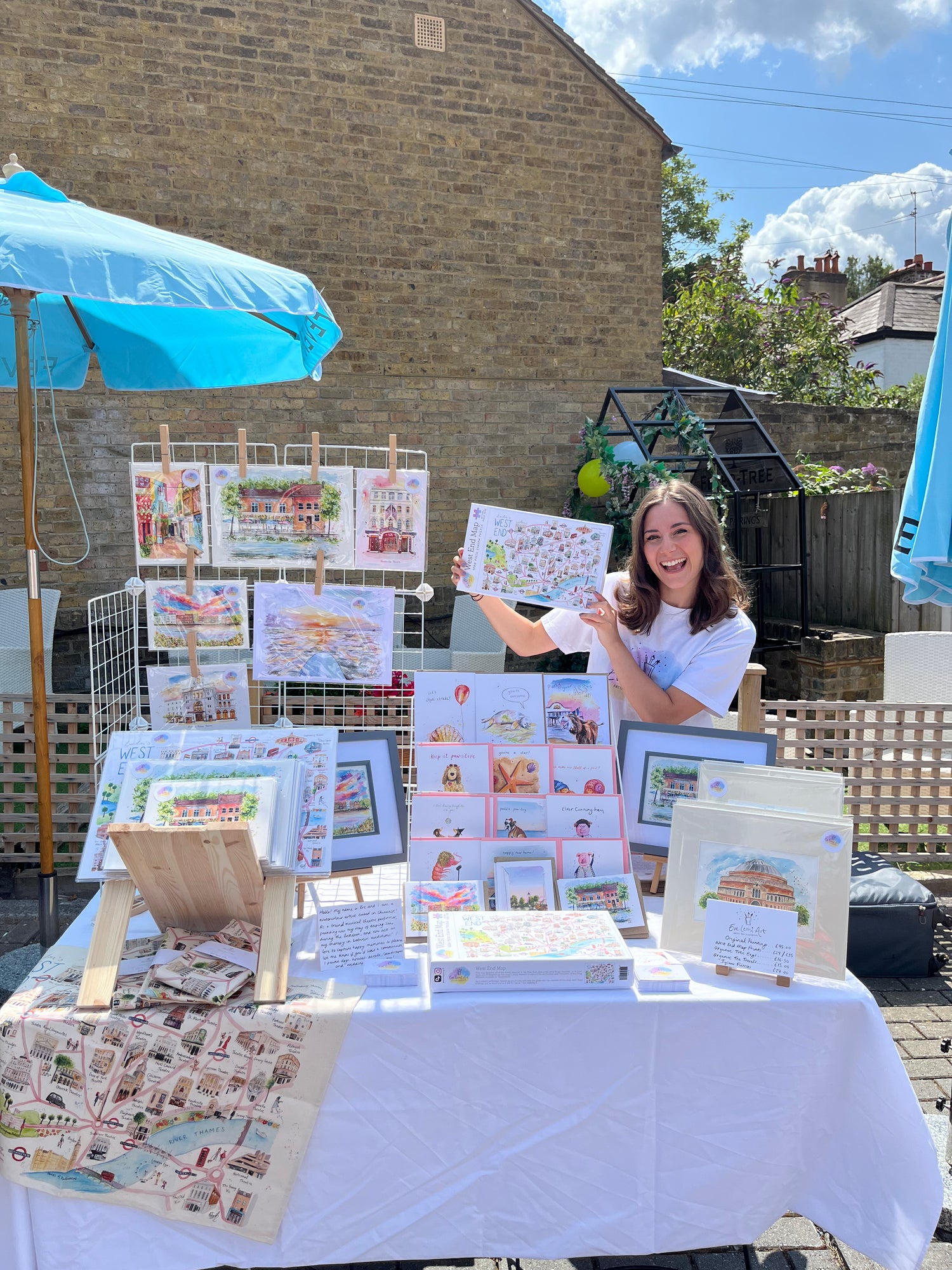 London artist Eve Leoni Smith holds up a West End Theatre Jigsaw Puzzle behind a table filled with illustrated prints, cards and musical theatre gifts.